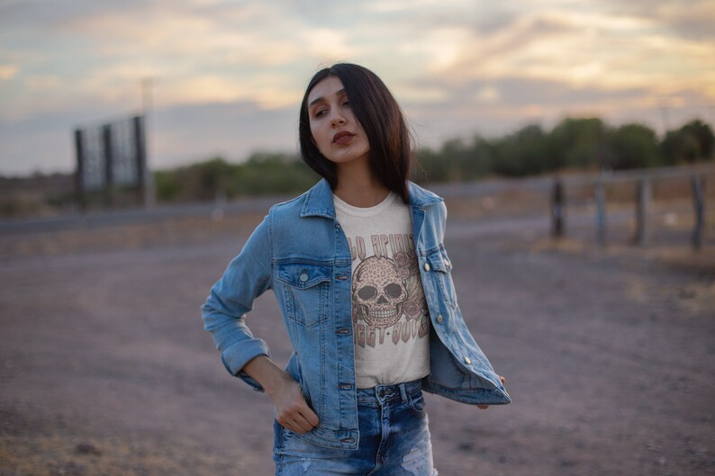 May include: A woman wearing a light blue denim jacket over a white t-shirt with a brown skull graphic and the text "Old Friends Get It Done".