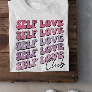 May include: White t-shirt with the text "SELF LOVE" repeated five times in a gradient of pink and purple. The text "Club" is written in cursive below the repeated text.