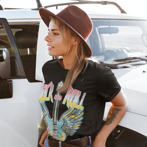 May include: A woman wearing a black t-shirt with a vintage-style graphic of a guitar with wings and the text "Rock and Roll". She is also wearing a brown hat, blue jeans, and a brown belt.