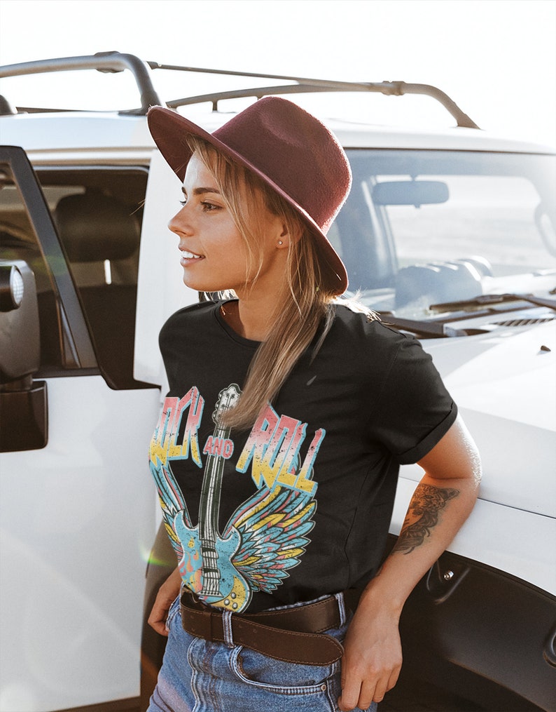 May include: A woman wearing a black t-shirt with a vintage-style graphic of a guitar with wings and the text "Rock and Roll". She is also wearing a brown hat, blue jeans, and a brown belt.