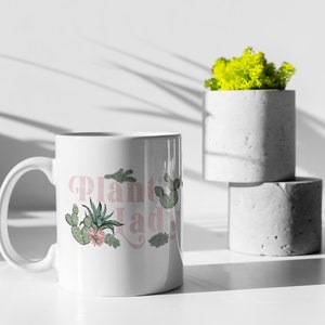 May include: White ceramic mug with a pink and green design that says "Plant Lady". The mug is sitting on a white surface next to two concrete planters.