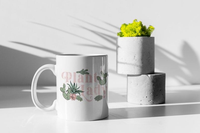 May include: White ceramic mug with a pink and green design that says "Plant Lady". The mug is sitting on a white surface next to two concrete planters.