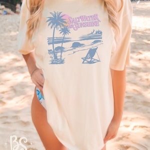 May include: A white t-shirt with a blue and pink graphic design of palm trees, a beach scene, and the text "Saltwater and Sunshine".