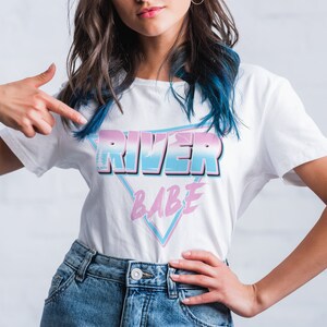 May include: A white t-shirt with a pink, blue, and white graphic design that says "River Babe" in a retro style.