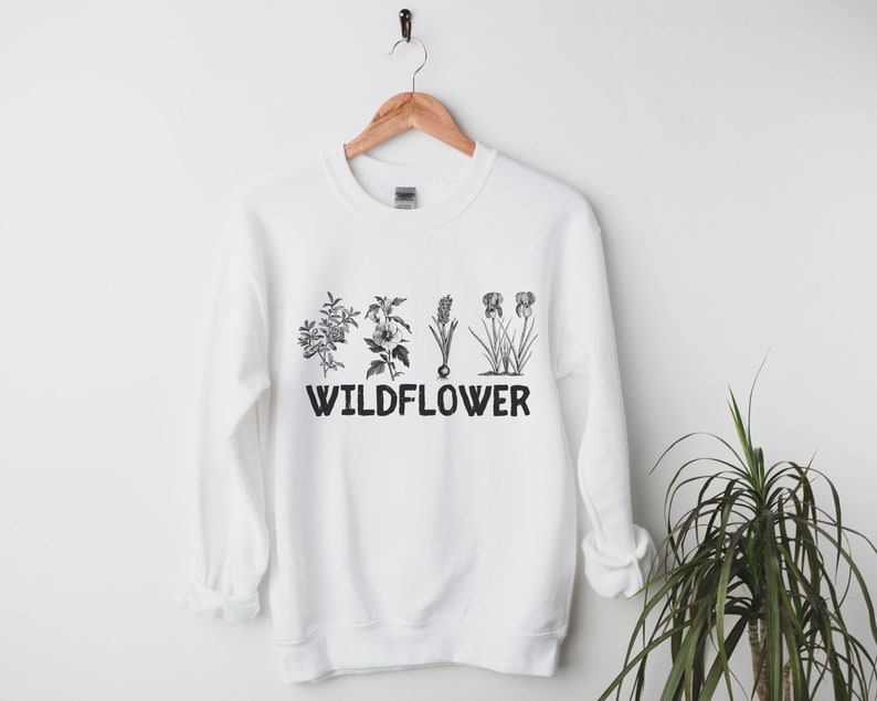 Pu&ograve; includere: Felpa bianca con una stampa nera di cinque diversi fiori selvatici e la parola "WILD FLOWER" sotto.