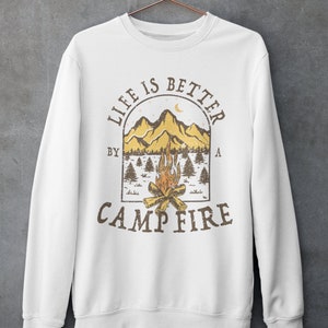 Pu&ograve; includere: Felpa bianca con una stampa grafica marrone di una scena di montagna con un fal&ograve; e il testo "Life is better by a campfire".