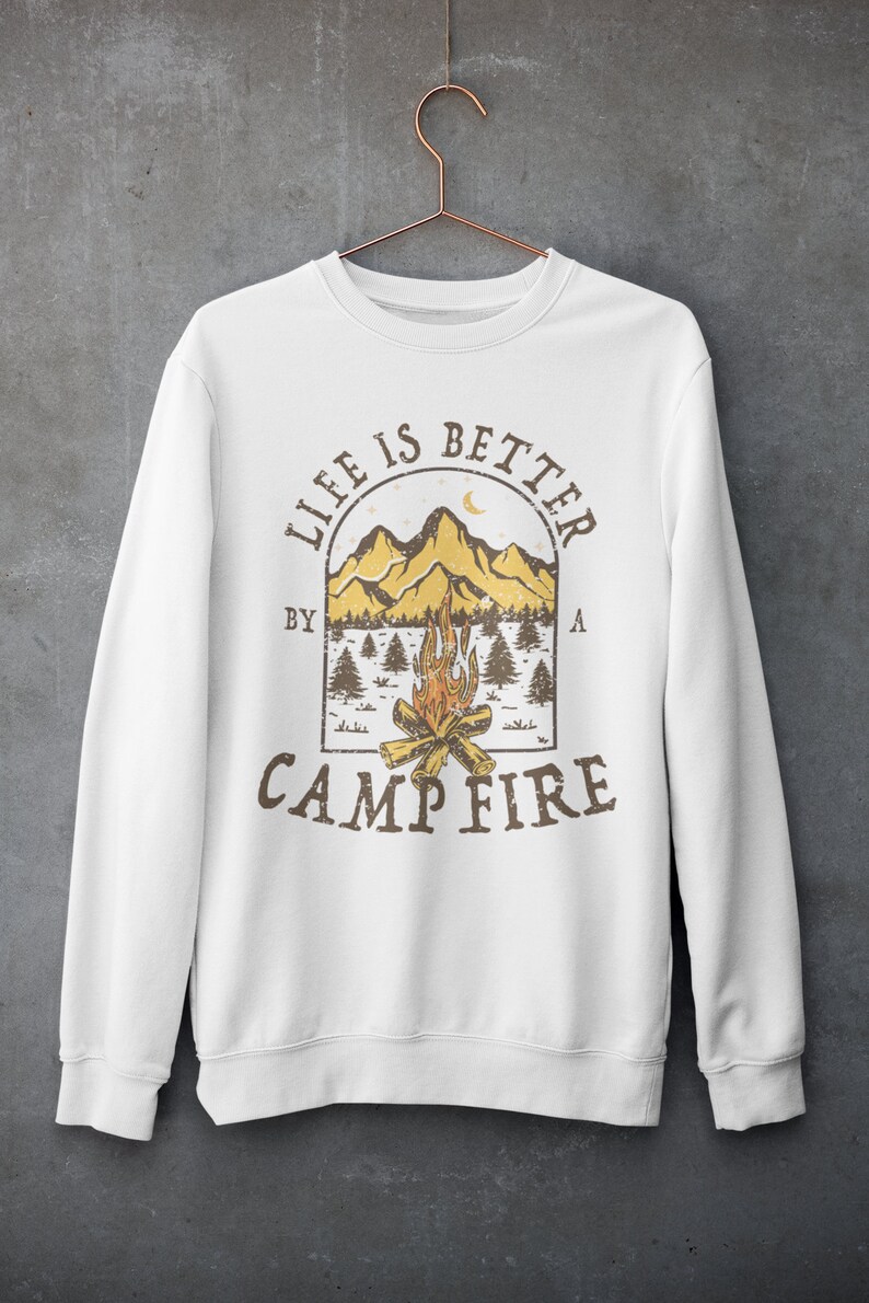 Pu&ograve; includere: Felpa bianca con una stampa grafica marrone di una scena di montagna con un fal&ograve; e il testo "Life is better by a campfire".