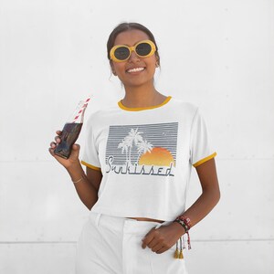 May include: A white cropped t-shirt with a yellow trim around the neckline and sleeves. The shirt features a graphic of palm trees and a sunset with the text "Sunkissed". The model is wearing white pants and yellow sunglasses.
