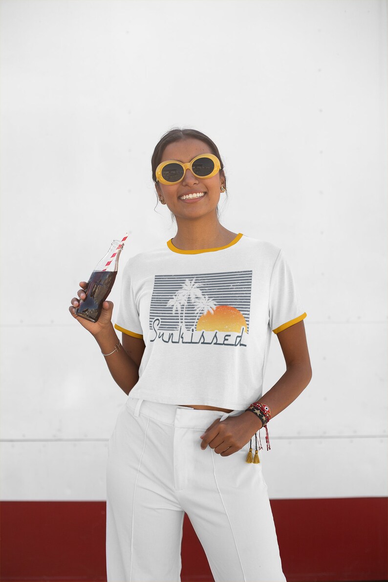 May include: A white cropped t-shirt with a yellow trim around the neckline and sleeves. The shirt features a graphic of palm trees and a sunset with the text "Sunkissed". The model is wearing white pants and yellow sunglasses.