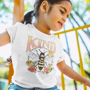 May include: A white t-shirt with a graphic of a bee and flowers. The text "KIND" is printed on the shirt in a rainbow gradient.