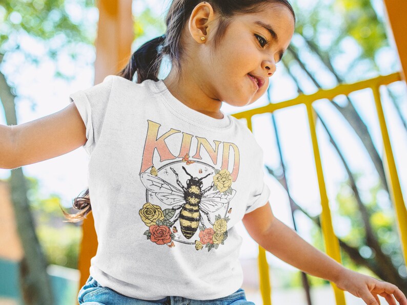 May include: A white t-shirt with a graphic of a bee and flowers. The text "KIND" is printed on the shirt in a rainbow gradient.