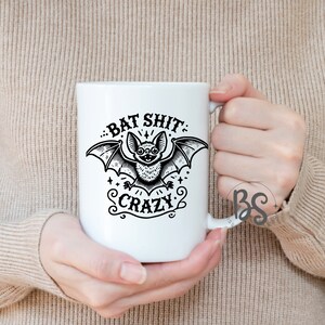 K&ouml;nnte beinhalten: Wei&szlig;e Keramiktasse mit einer schwarz-wei&szlig;en Illustration einer Fledermaus mit dem aufgedruckten Text "Bat Shit Crazy".