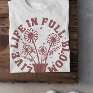 Op de afbeelding: Wit T-shirt met een bruine afbeelding van bloemen in een pot en de tekst "Live Life in Full Bloom" in een cirkel rond de bloemen.