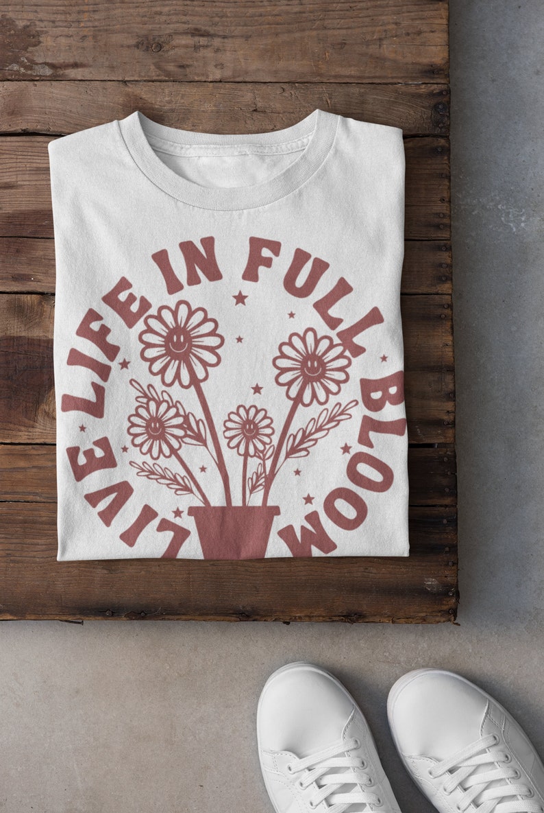 Op de afbeelding: Wit T-shirt met een bruine afbeelding van bloemen in een pot en de tekst "Live Life in Full Bloom" in een cirkel rond de bloemen.