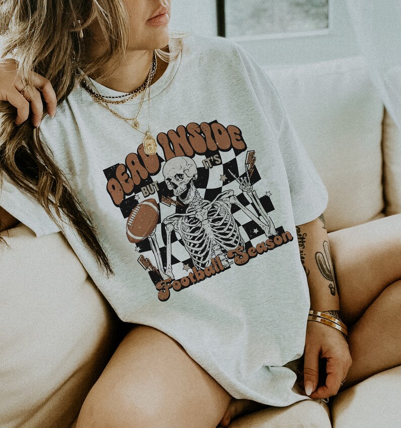 Puede incluir: Una camiseta blanca con un gr&aacute;fico de un esqueleto sosteniendo un bal&oacute;n de f&uacute;tbol y el texto "Dead Inside But It's Football Season".