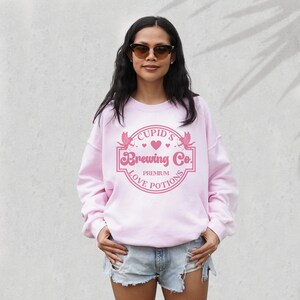 May include: Pink sweatshirt with a pink graphic of a heart with wings and the text "Cupid's Brewing Co. Premium Love Potions"