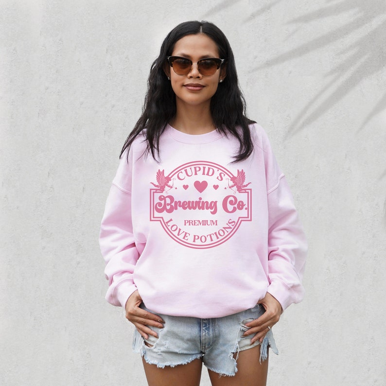 May include: Pink sweatshirt with a pink graphic of a heart with wings and the text "Cupid's Brewing Co. Premium Love Potions"