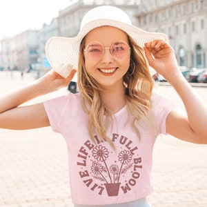 Op de afbeelding: Een roze T-shirt met een afbeelding van een bloempot met drie bloemen en de tekst "Live Life in Full Bloom".