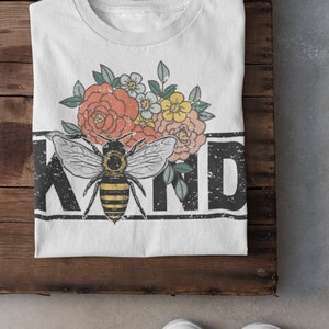 Pu&ograve; includere: Maglietta bianca con una grafica in bianco e nero di un'ape e fiori. Il testo "KIND" &egrave; stampato in nero con un effetto vintage.