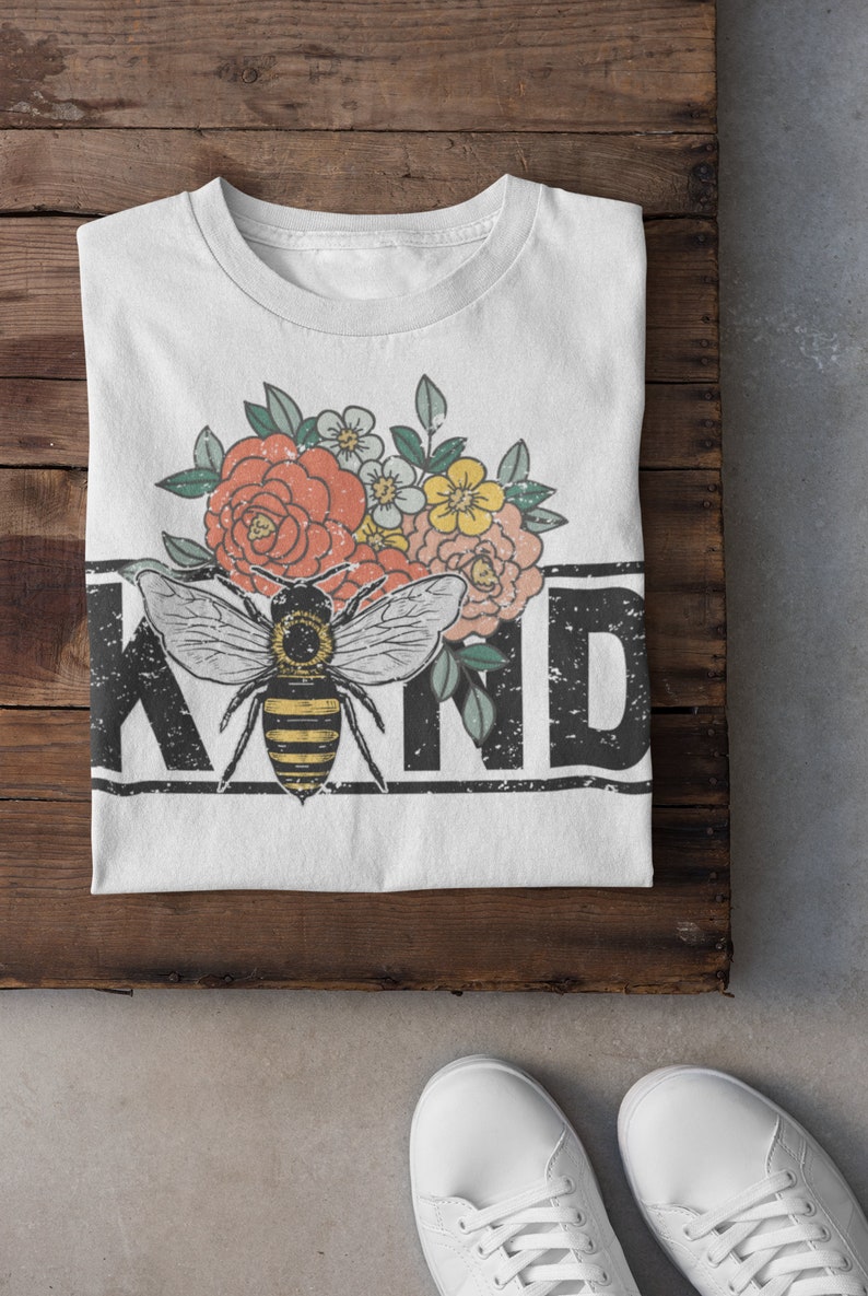 Pu&ograve; includere: Maglietta bianca con una grafica in bianco e nero di un'ape e fiori. Il testo "KIND" &egrave; stampato in nero con un effetto vintage.