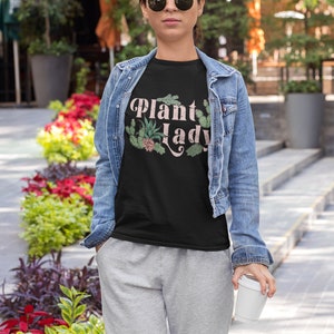 May include: A woman wearing a black t-shirt with the text "Plant Lady" in pink lettering and a green plant graphic. She is wearing a denim jacket and grey sweatpants.