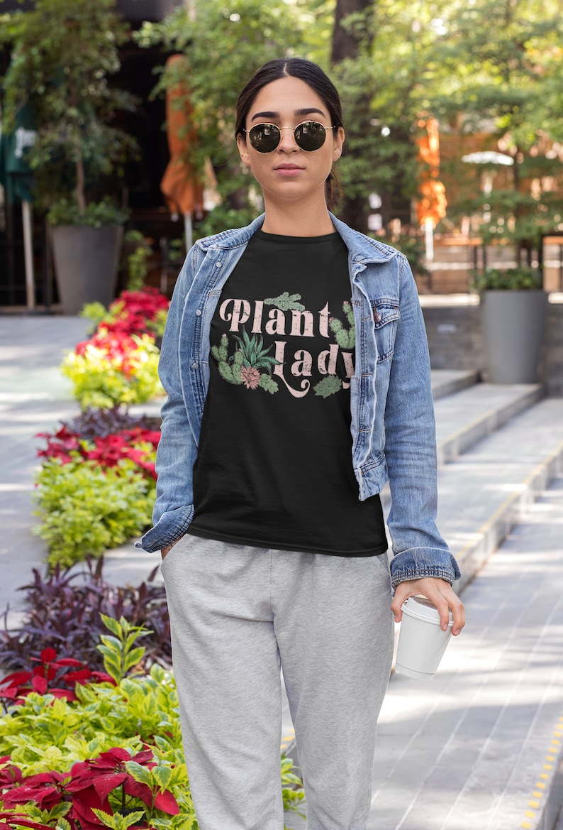 May include: A woman wearing a black t-shirt with the text "Plant Lady" in pink lettering and a green plant graphic. She is wearing a denim jacket and grey sweatpants.