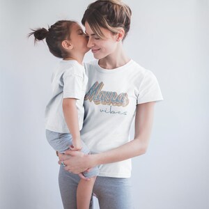 Puede incluir: Una mujer y una ni&ntilde;a llevan camisetas blancas. La camiseta de la mujer tiene el texto "Mama vibes" en una fuente retro colorida. La ni&ntilde;a est&aacute; besando a la mujer en la mejilla.