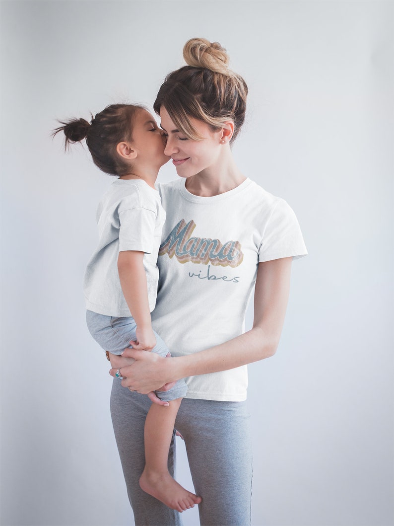 Puede incluir: Una mujer y una ni&ntilde;a llevan camisetas blancas. La camiseta de la mujer tiene el texto "Mama vibes" en una fuente retro colorida. La ni&ntilde;a est&aacute; besando a la mujer en la mejilla.