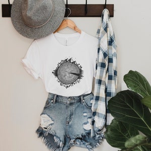 May include: White t-shirt with a black and white graphic of a tree ring with animals and plants around it. The shirt is hanging on a wooden hanger with a gray hat and a blue and white plaid shirt hanging next to it.
