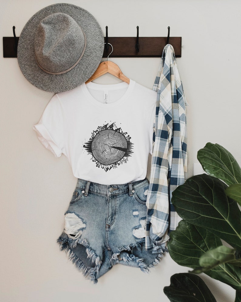 May include: White t-shirt with a black and white graphic of a tree ring with animals and plants around it. The shirt is hanging on a wooden hanger with a gray hat and a blue and white plaid shirt hanging next to it.
