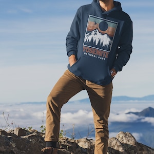 May include: A navy blue hoodie featuring a vintage-style graphic of a mountain range with a sun setting behind it. The graphic also includes the text "Yosemite National Park".