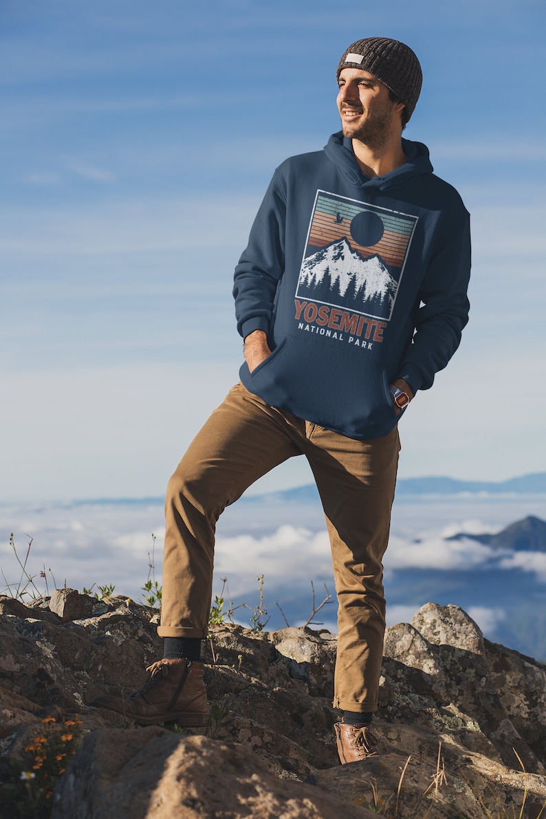 May include: A navy blue hoodie featuring a vintage-style graphic of a mountain range with a sun setting behind it. The graphic also includes the text "Yosemite National Park".