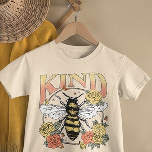 May include: A beige t-shirt with a graphic of a bee surrounded by flowers and the word "KIND" in a vintage style font.