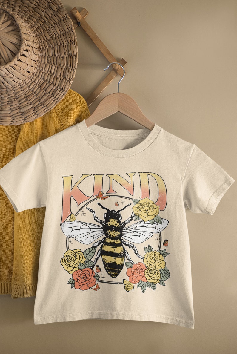 May include: A beige t-shirt with a graphic of a bee surrounded by flowers and the word "KIND" in a vintage style font.
