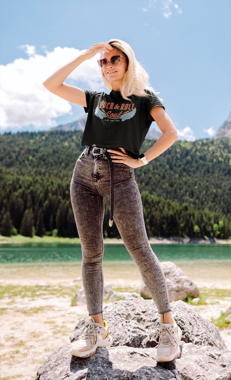 Puede incluir: Una mujer con una camiseta negra con el texto "Rock & Roll Soul" y alas, jeans grises y zapatillas blancas. Est&aacute; de pie sobre una roca junto a un lago.
