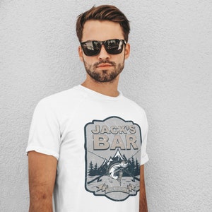 May include: White t-shirt with a vintage-style logo that says "Jack's Bar" with a mountain scene and a fish. The logo also says "Virgin River, CA".