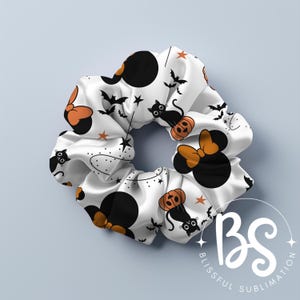 May include: White scrunchie with a Halloween-themed print featuring black cats, pumpkins, and Minnie Mouse ears. The print is repeated throughout the scrunchie.
