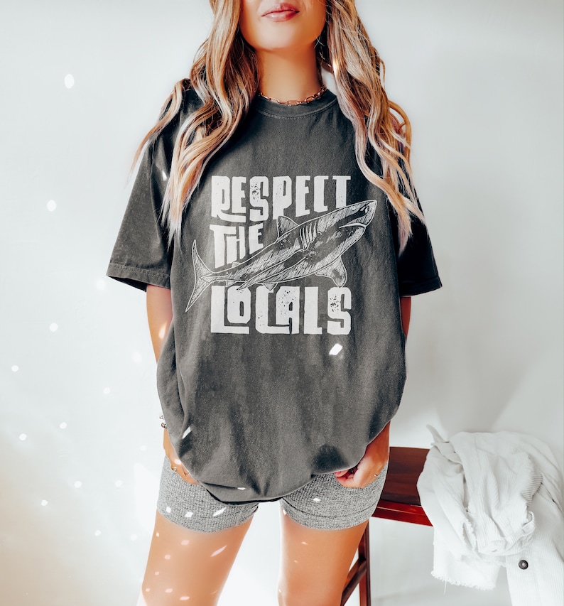a woman wearing an oversize sweatshirt shirt.  the shirt features a graphic of a shark and the words "respect the locals" in a retro text.  she is standing in front of a white wall.