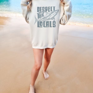 a woman wearing an oversize sweatshirt shirt.  the shirt features a graphic of a shark and the words "respect the locals" in a retro text.  she is standing in front of the ocean