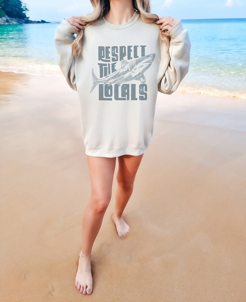 a woman wearing an oversize sweatshirt shirt.  the shirt features a graphic of a shark and the words "respect the locals" in a retro text.  she is standing in front of the ocean