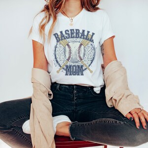 Op de afbeelding: Wit T-shirt met een blauwe en witte baseball afbeelding en de tekst "Baseball Mom".