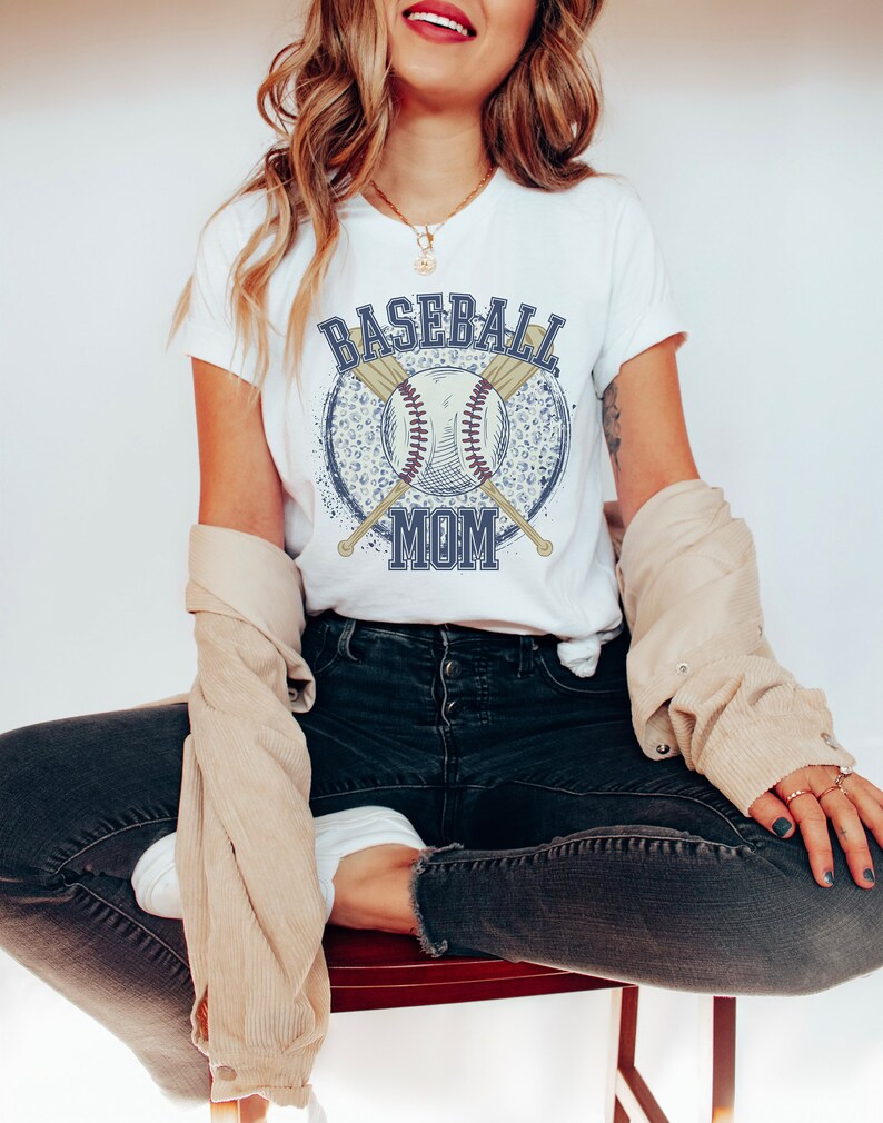 Op de afbeelding: Wit T-shirt met een blauwe en witte baseball afbeelding en de tekst "Baseball Mom".