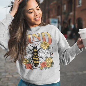 May include: Gray sweatshirt with a bee and floral design. The text "KIND" is printed on the sweatshirt.