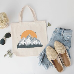 May include: A white canvas tote bag with a mountain range and a setting sun graphic. The bag is next to a pair of straw sandals and a pair of blue denim shorts.