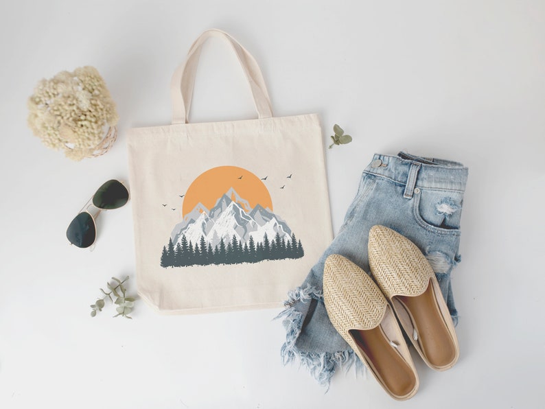 May include: A white canvas tote bag with a mountain range and a setting sun graphic. The bag is next to a pair of straw sandals and a pair of blue denim shorts.