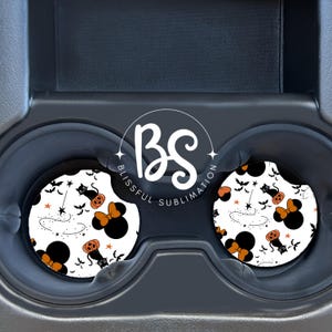 May include: Two white car coasters with a black and orange Halloween themed design featuring a black cat, a pumpkin, a magic wand, stars, and a Minnie Mouse head with a bow. The coasters are in a car cup holder.