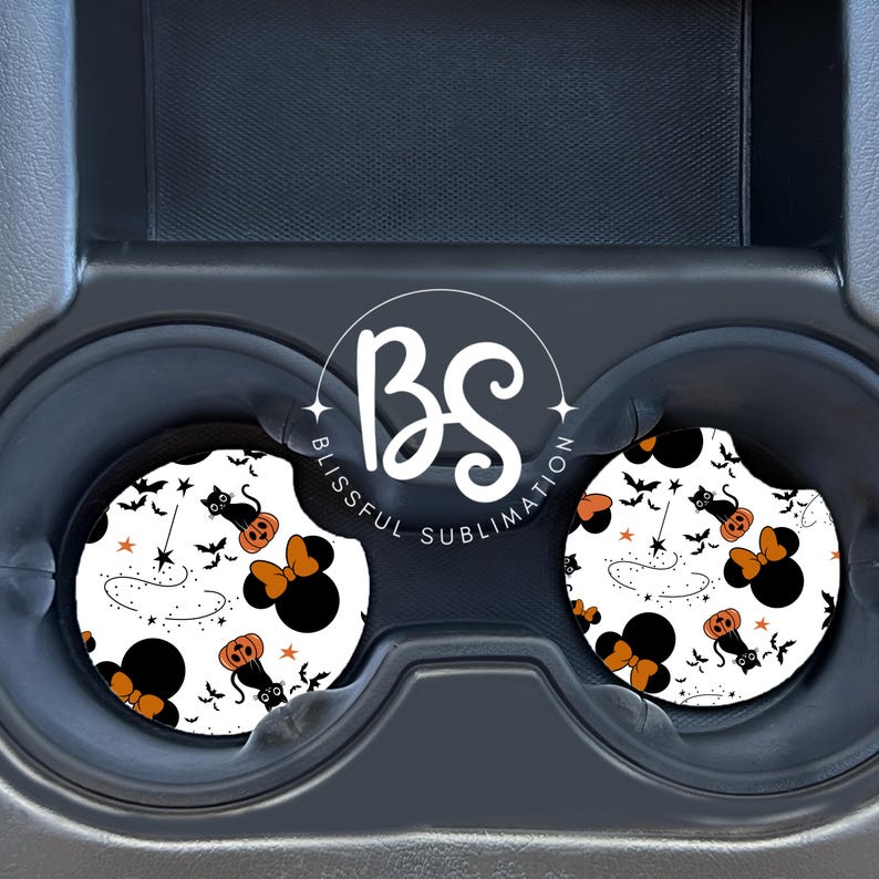 May include: Two white car coasters with a black and orange Halloween themed design featuring a black cat, a pumpkin, a magic wand, stars, and a Minnie Mouse head with a bow. The coasters are in a car cup holder.