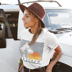 May include: White t-shirt with a graphic of palm trees and a sunset with the word "kissed" underneath. The model is wearing a brown hat and blue jeans.