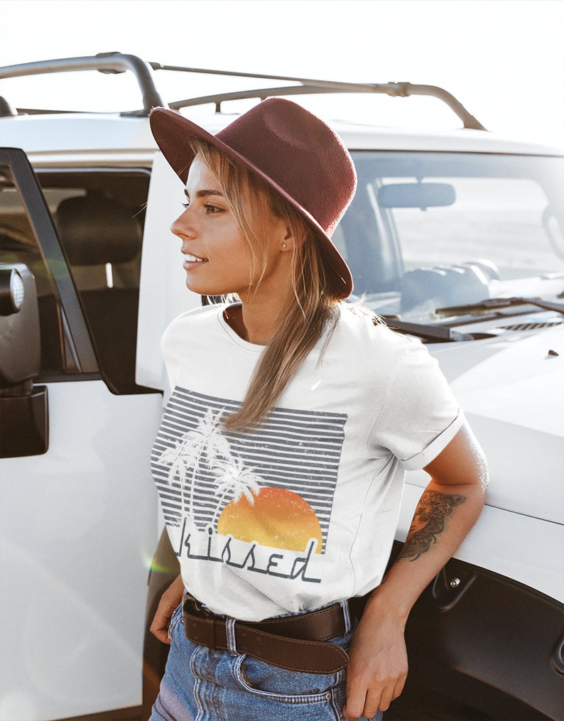 May include: White t-shirt with a graphic of palm trees and a sunset with the word "kissed" underneath. The model is wearing a brown hat and blue jeans.