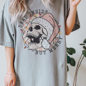 May include: A light blue t-shirt with a graphic of a skull wearing a Santa hat and surrounded by Christmas lights. The text "Dead Inside But Merry" is printed on the shirt.